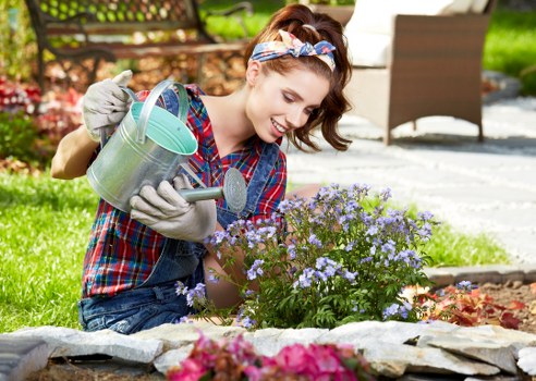 A young woman with light skin, brown hair tied back with a colorful headband, is smiling while watering a flowering shrub with purple blossoms in a well-maintained front garden. She is dressed in casual outdoor clothing, including a blue plaid shirt and gardening gloves. The garden features a neat stone-edged flower bed with blooming plants and soil, bordered by paving slabs and a lush green lawn. In the background, there are garden furniture pieces, such as a brown outdoor chair, and a wooden fence, indicating an inviting outdoor space. The scene is set during daylight with clear weather, highlighting the vibrant natural colours of the plants, grass, and garden features, which aligns with gardening and landscaping services available in Camberwell and the surrounding areas.