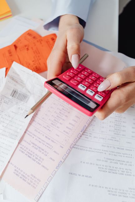A close-up of a person's hand holding a pink calculator and using it on a cluttered desk with various documents, receipts, and a pencil. The documents appear to be related to financial or gardening se